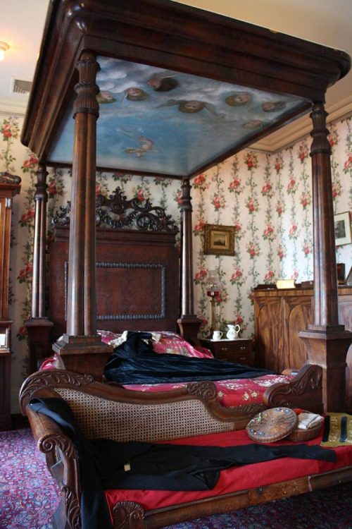 A vintage four-poster bed with a painted ceiling, surrounded by floral wallpaper and antique furniture.