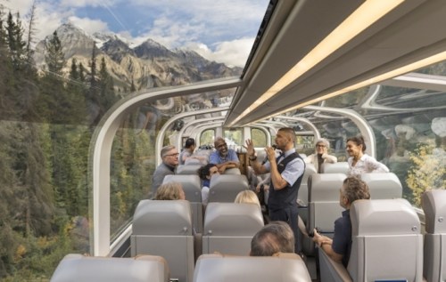 A scenic train interior with passengers enjoying views of mountains through large windows. A staff member is speaking.