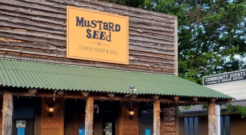 Exterior of Mustard Seed Coffee Shop & Deli, featuring a wooden facade and a green roof, with a community events sign nearby.