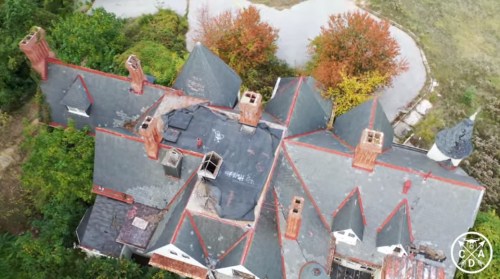 Aerial view of a large, abandoned house with a complex roof and autumn trees in the background.