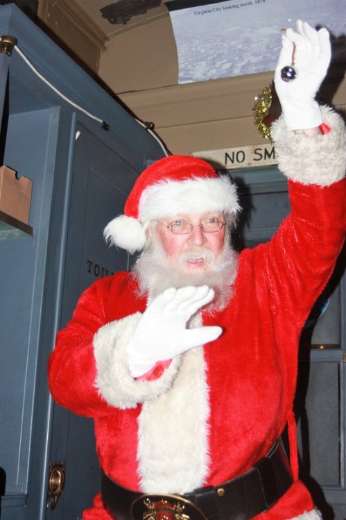 A cheerful Santa Claus in a red suit and white gloves waves joyfully, standing in a festive indoor setting.