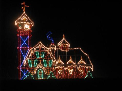 A brightly lit holiday display featuring a whimsical house and tower, adorned with colorful lights against a dark background.