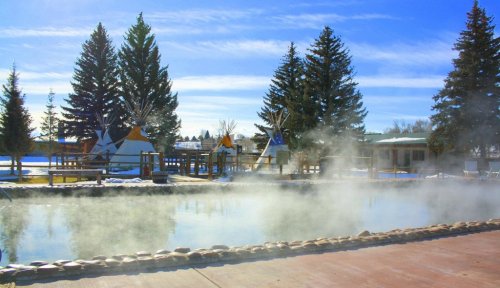 A serene outdoor scene featuring a steaming hot spring surrounded by teepees and tall pine trees under a clear blue sky.