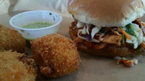 A close-up of a pulled chicken sandwich with coleslaw, accompanied by crispy fried balls and a small cup of green sauce.