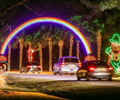 Colorful holiday lights form a rainbow arch over a road, with festive decorations and cars passing by.