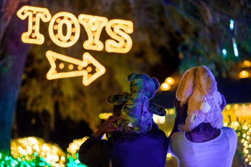 Two children wearing animal hats look at a glowing "TOYS" sign with an arrow, surrounded by festive lights.