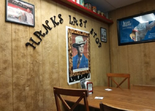 A rustic diner interior with wooden walls, a framed cowboy portrait, and the words "Hank's Last Stop" on the wall.