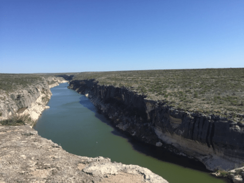 A serene river flows through a canyon, surrounded by rocky cliffs and green vegetation under a clear blue sky.