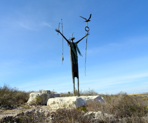 A tall bronze statue of a figure with outstretched arms, holding a bird and objects, set against a blue sky.