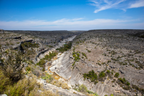 A vast canyon landscape with rocky cliffs, sparse vegetation, and a winding river under a clear blue sky.