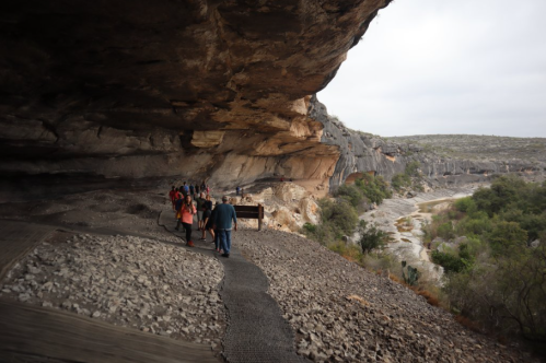 Visitors walk along a rocky path under a large overhang, with a scenic view of a river and hills in the background.