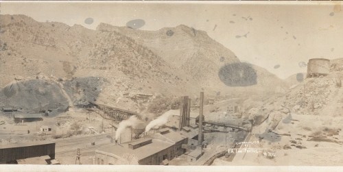 A sepia-toned landscape of a mining site with buildings, smokestacks, and mountains in the background.