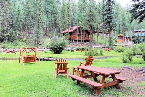 A serene forest scene featuring wooden chairs and tables, surrounded by trees and a cozy cabin in the background.