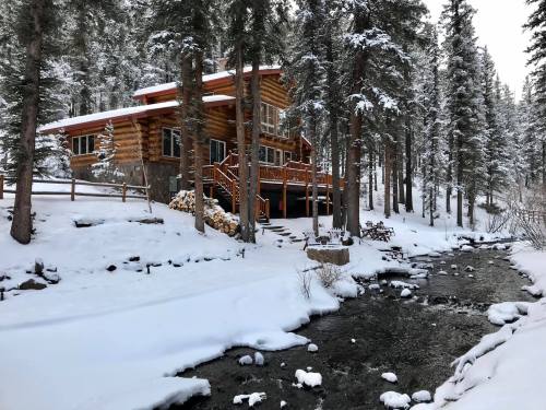 A cozy log cabin surrounded by snow-covered trees, next to a flowing stream in a winter landscape.