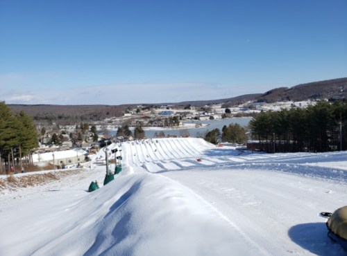 A snowy landscape with tubing hills, trees, and a distant view of a town and lake under a clear blue sky.