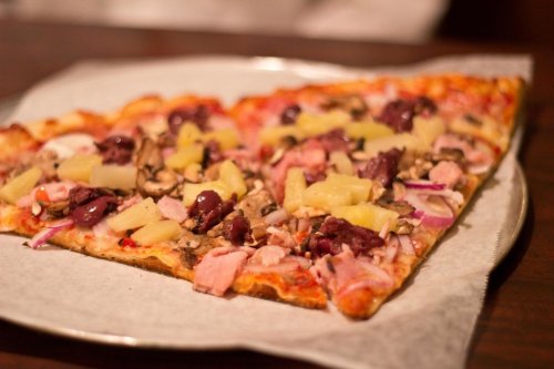 A slice of pizza topped with ham, pineapple, olives, mushrooms, and red onions on a paper-lined plate.