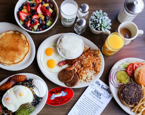 A table spread with breakfast items: pancakes, eggs, bacon, hash browns, fruit, juice, and a burger with toppings.
