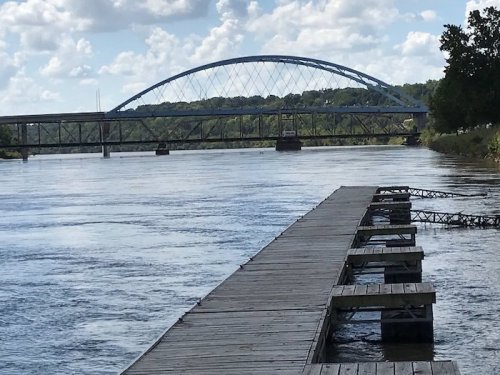 A wooden dock extends into a river, with a large arch bridge spanning the water in the background under a cloudy sky.