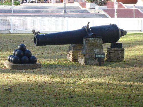 A historic cannon on display in a grassy area, with a stack of cannonballs nearby and a sign indicating its history.