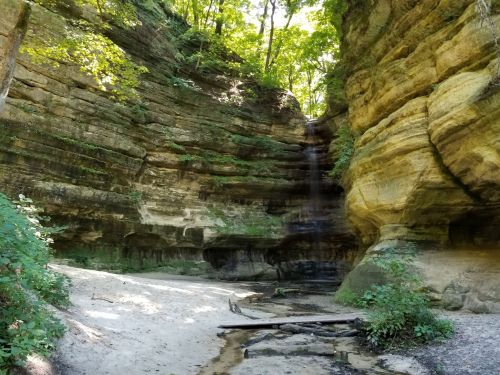 A serene waterfall cascading down rocky cliffs, surrounded by lush greenery and sandy ground in a tranquil natural setting.