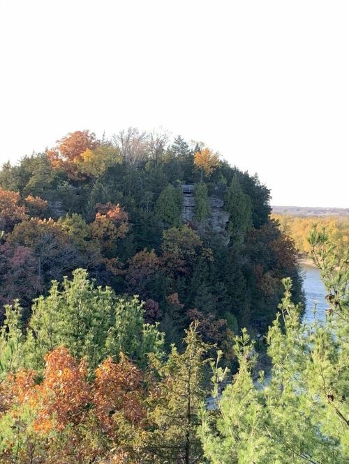 A scenic view of a rocky cliff surrounded by colorful autumn trees and a river in the background.