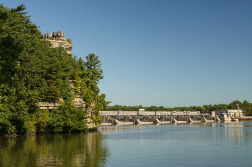 A scenic view of a dam beside a calm river, with a rocky cliff and trees in the foreground under a clear blue sky.