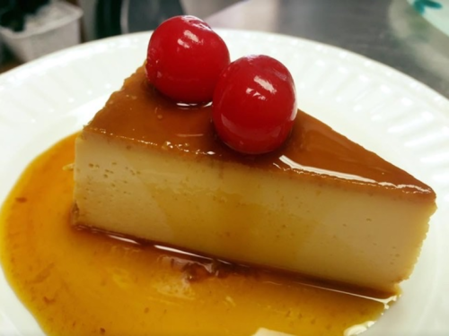 A slice of flan topped with two red cherries, served on a white plate with caramel sauce.