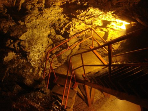 A dimly lit underground staircase with metal railings, surrounded by rocky walls and warm lighting.