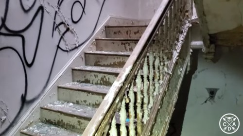 A worn staircase with peeling paint and graffiti on the wall, leading to an upper level in a dilapidated building.