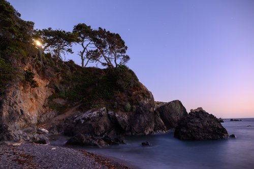 A serene coastal scene at dusk, featuring rocky cliffs, gentle waves, and trees silhouetted against a twilight sky.