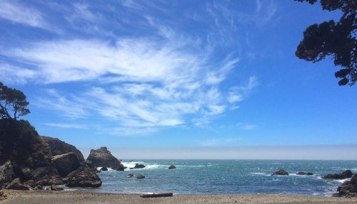 A serene beach scene with rocky cliffs, gentle waves, and a bright blue sky with wispy clouds.