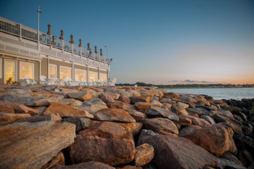 A rocky shoreline at sunset with a building featuring large windows and outdoor seating overlooking the water.