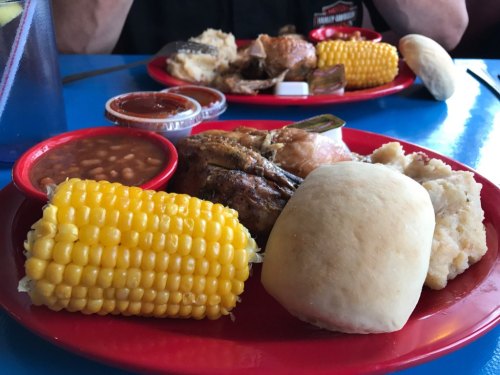 A colorful plate featuring grilled chicken, corn on the cob, baked beans, a roll, and a side of sauce.