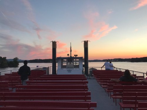 A sunset view from a ferry with red benches, featuring a calm lake and colorful clouds in the sky.