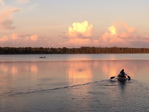 A serene lake at sunset, with a person rowing a small boat and soft clouds reflecting on the water's surface.