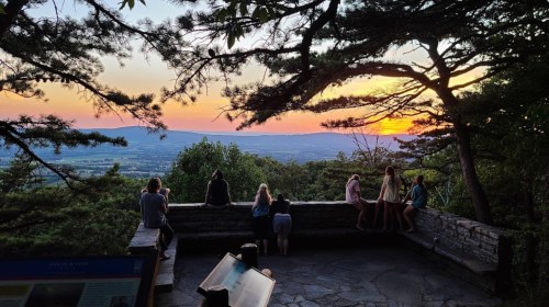 People sit on a stone wall, watching a colorful sunset over a valley, framed by trees.