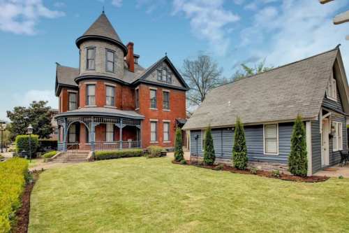 A large red Victorian house with a turret, alongside a smaller gray house, set in a well-maintained lawn.