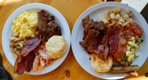 Two plates of hearty breakfast: scrambled eggs, bacon, biscuits, potatoes, and various meats.