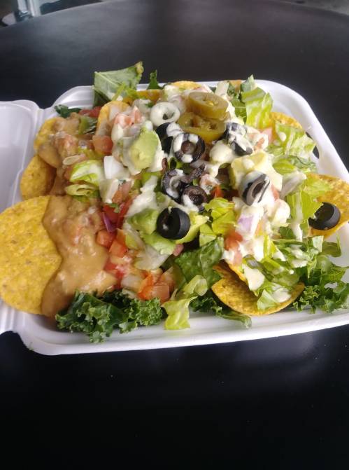 A takeout container filled with nachos topped with beans, lettuce, tomatoes, olives, jalapeños, and sour cream.