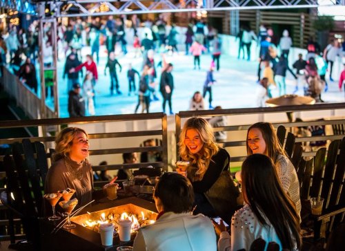A group of friends laughing around a fire pit, with an ice skating rink and crowd in the background.