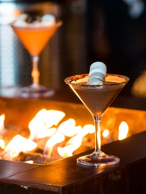 A chocolate martini with a marshmallow garnish sits in front of a warm, glowing fire. Another cocktail is blurred in the background.