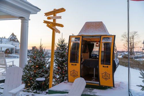 A yellow cabin with open doors sits in the snow, surrounded by Christmas trees and a directional signpost.
