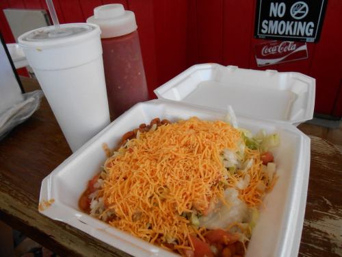 A takeout container filled with a salad topped with shredded cheese, alongside a drink and sauce in the background.