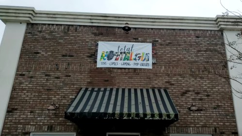 A brick building with a sign reading "Total" and colorful text below it, advertising toys, comics, gaming, and pop culture.