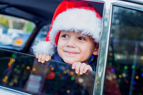 A smiling child in a Santa hat leans out of a car window, surrounded by colorful holiday lights.