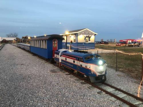 A vintage-style train with a blue and orange engine parked beside a blue train station at dusk.