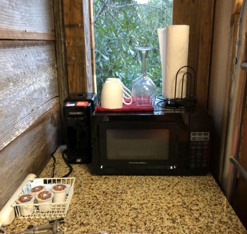 A countertop with a microwave, coffee maker, cups, and paper towels, set against a rustic wooden wall.