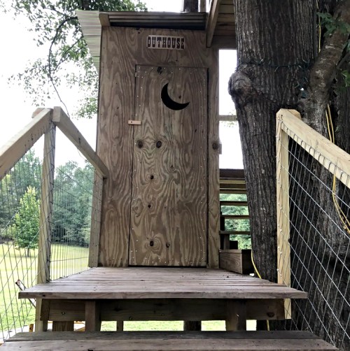 A wooden outhouse with a crescent moon cutout door, situated on a raised platform among trees.