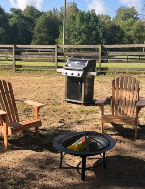 A backyard scene featuring two wooden chairs, a fire pit, and a grill surrounded by a grassy area and wooden fence.
