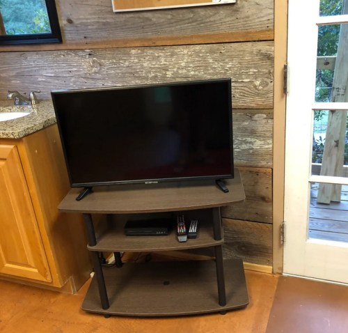 A flat-screen TV on a wooden stand, placed against a rustic wall with a door and kitchen counter nearby.